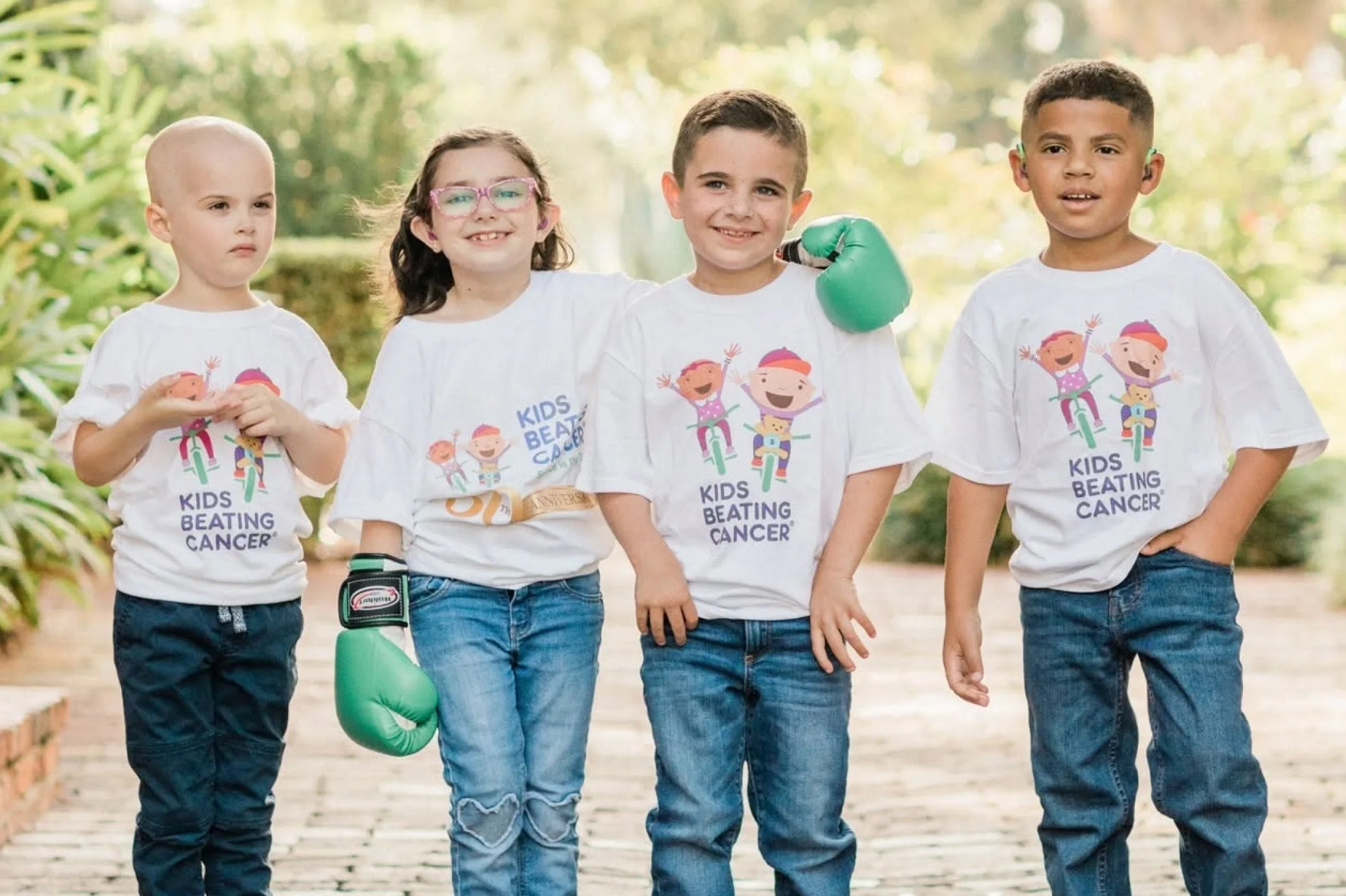 Four young children standing outdoors together wearing white "Kids Beating Cancer" T-shirts and green boxing gloves.