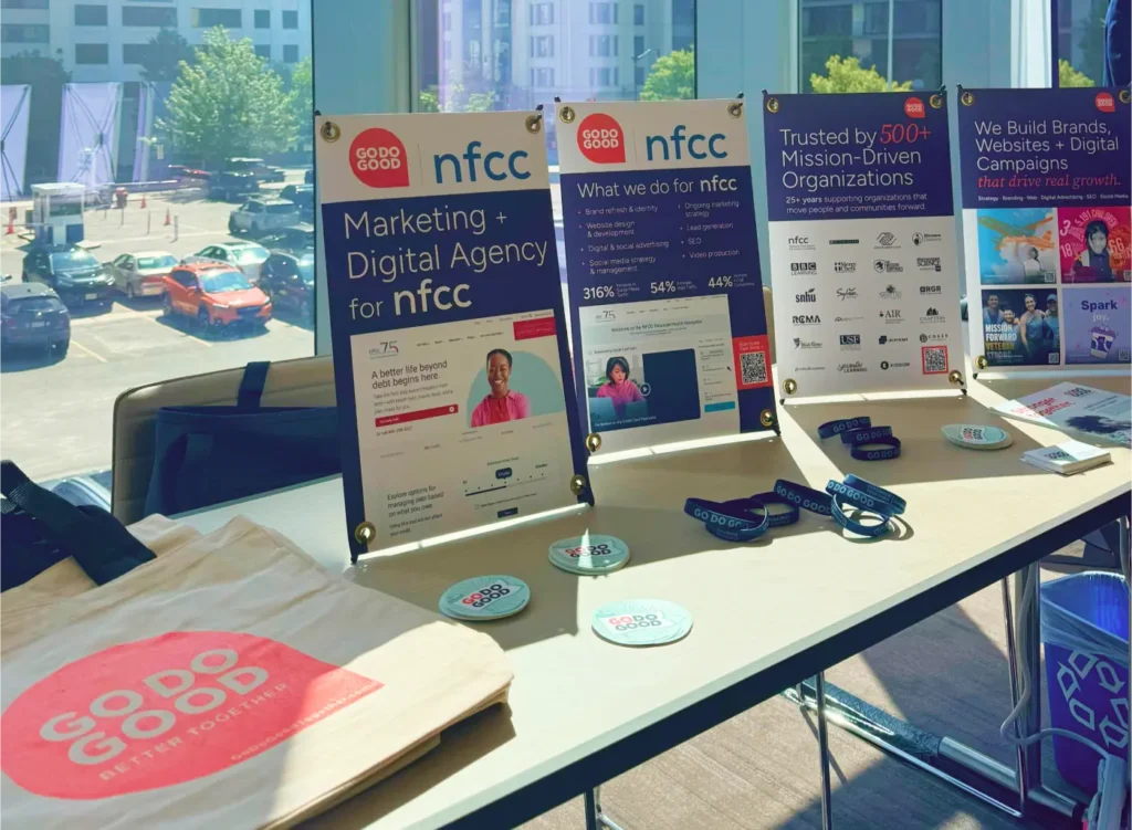 A sponsor vendor table situated by a window, displaying NFCC signage, promotional materials, and a pink Go Do Good branded tote bag.