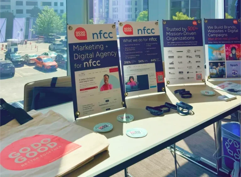 A sponsor vendor table situated by a window, displaying NFCC signage, promotional materials, and a pink Go Do Good branded tote bag.