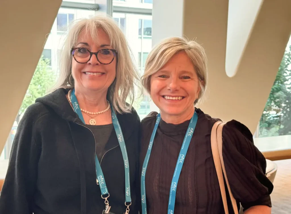 Jess and Michelle smiling together inside the bright conference venue, both wearing blue NFCC Connect attendee lanyards.