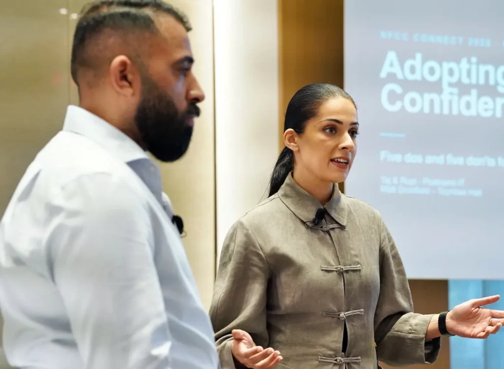 Two speakers, a man and a woman, presenting on stage at the NFCC Connect conference with a background slide titled "Adopting AI with Confidence."