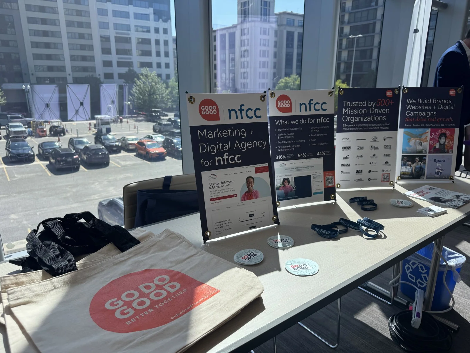 A conference exhibition table featuring Go Do Good and NFCC promotional banners, tote bags, branded wristbands, and stickers by a large window overlooking a parking lot.