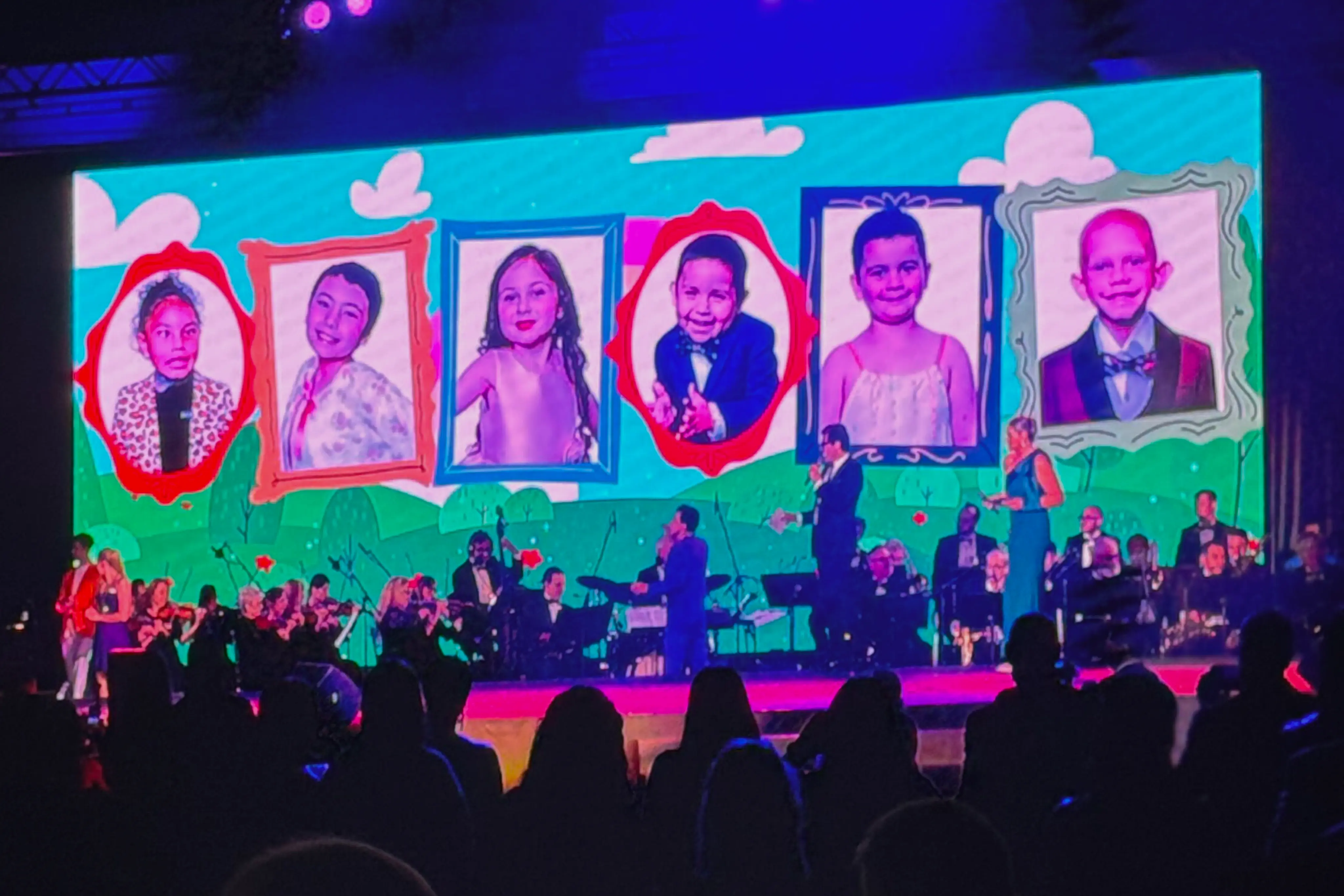 Stage screen displaying portraits of children in colorful frames above a live orchestra performing for a seated audience at a gala.